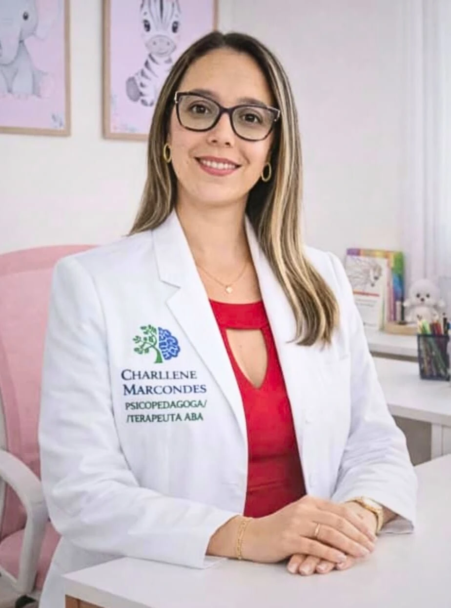 Sou uma mulher adulta de pele clara, com cabelos longos castanho-claros com mechas loiras, lisos e soltos. Uso óculos de armação preta e brincos pequenos dourados. Estou sorrindo levemente e olhando para a câmera. Visto blusa vermelha e jaleco branco com a logo da clínica (a mesma do site).
                Estou sentada atrás de uma mesa clara, com as mãos apoiadas uma sobre a outra. Ao fundo, meu consultório tem decoração suave em tons claros, com quadros infantis na parede, estante com livros e objetos e iluminação acolhedora.