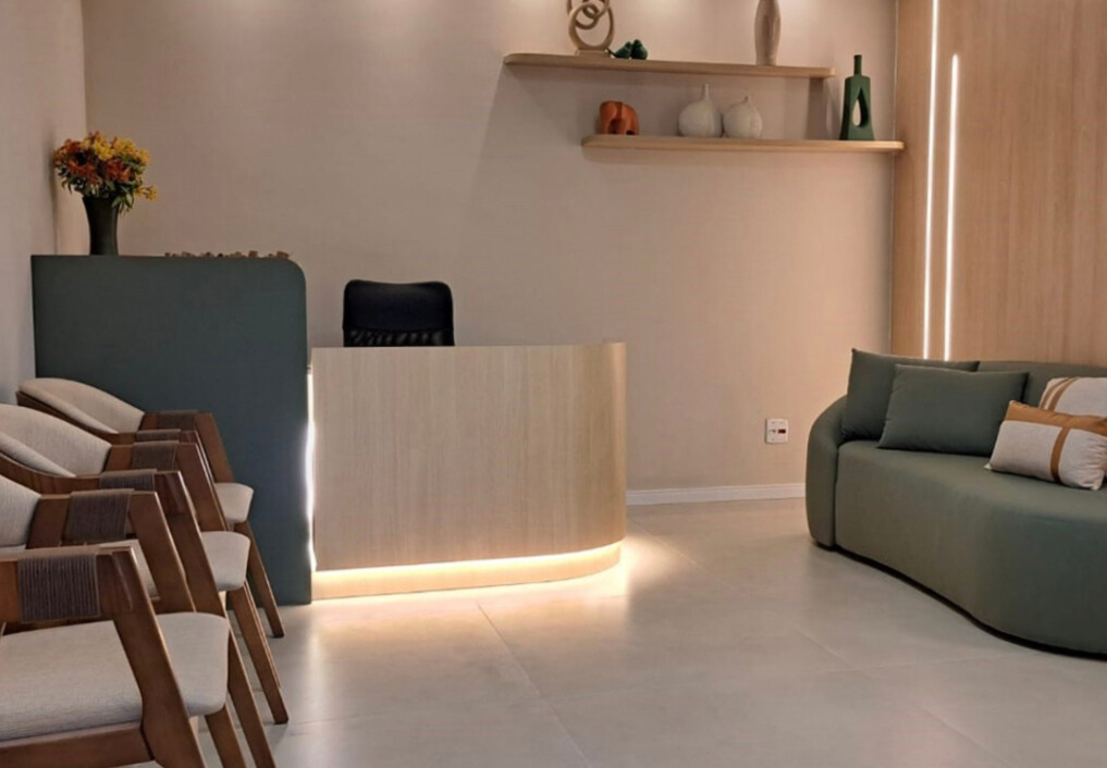 Recepção com balcão de madeira iluminado, cadeiras alinhadas, sofá verde e decoração minimalista.