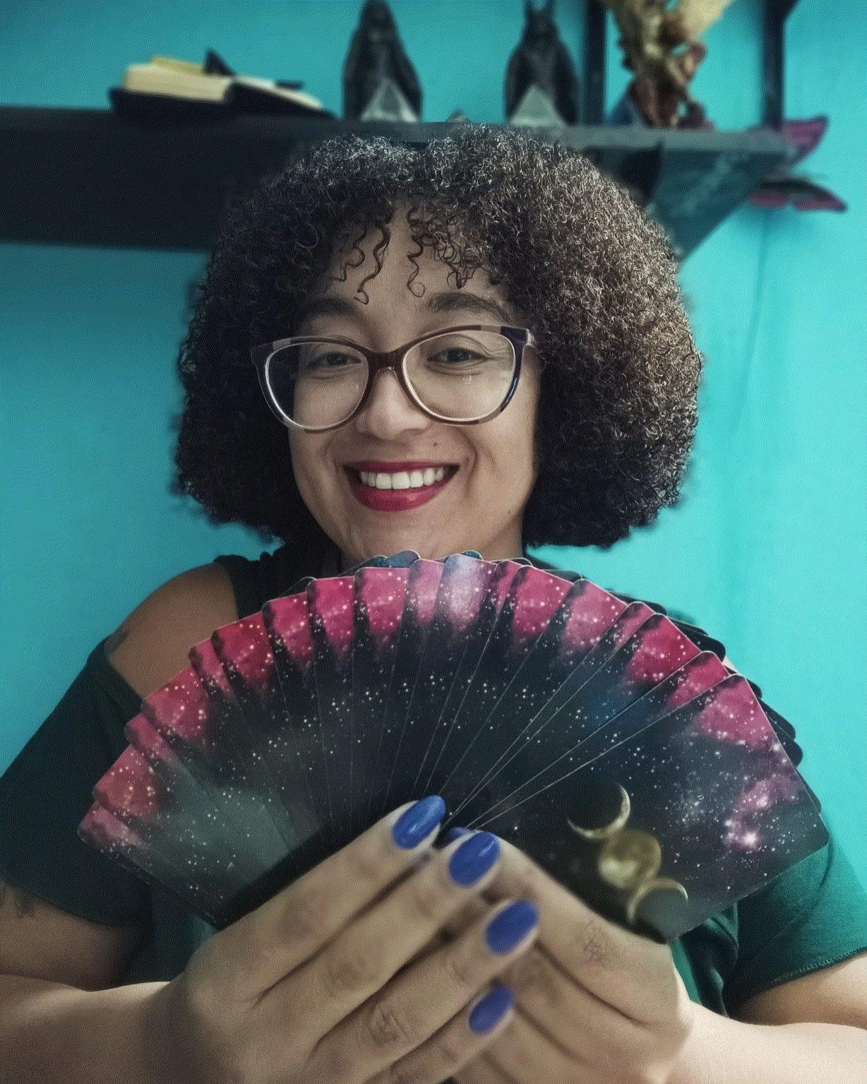 foto de Jaqueline: Negra, cabelos cacheados, usando óculos, sorrindo e segurando cartas de Trot em suas mãos.